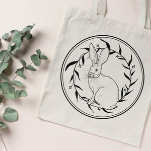 May include: A white tote bag with a black and white illustration of a rabbit inside a wreath of leaves. The rabbit is sitting upright with its ears perked up.