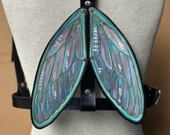 Puede incluir: Un arnés de cuero negro con dos alas grandes de color verde iridiscente unidas. Las alas tienen un diseño pintado detallado.