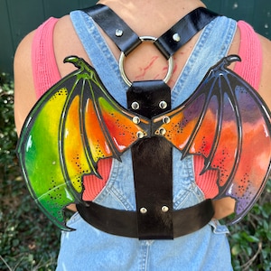 May include: A black leather harness with two rainbow-coloured dragon wings attached. The wings are painted with green, yellow, orange, red, and purple.