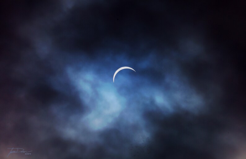 Op de afbeelding: Een sikkelvormige zonsverduistering zichtbaar door een bewolkte lucht. De verduistering is een dunne strook licht, omringd door donkere wolken.