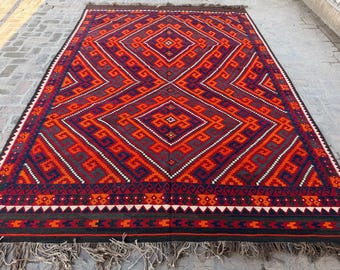 May include: A rectangular, hand-woven rug with a geometric diamond pattern. The rug features a central design in shades of red, orange, and navy blue, framed by a dark border and fringed edges. The rug's dimensions are not visible.