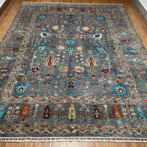 May include: A large, rectangular area rug with a gray background and an intricate floral pattern in shades of teal, red, orange, and yellow. The rug has a detailed border and a fringe along the edges, suitable for a living room or bedroom.