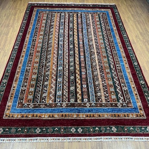 May include: A rectangular area rug with a striped pattern in shades of red, blue, brown, and gold. The rug has a dark red border with a green and white floral design. The central design features vertical stripes of varying widths and colors.