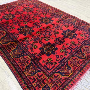 May include: A rectangular area rug featuring a vibrant red background with repeating dark blue geometric patterns and floral motifs. The rug includes a wide border with intricate dark blue and yellow designs and white fringe on the short ends.