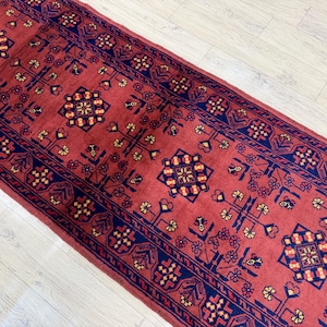 May include: A long rectangular runner rug featuring a deep red background with a repeating pattern of dark blue geometric medallions and stylized floral motifs in yellow, cream, and dark blue, arranged in a symmetrical layout across the surface.