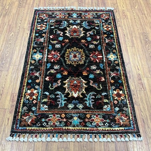 Könnte beinhalten: Ein rechteckiger, handgeknüpfter Teppich mit schwarzem Hintergrund und aufwendigen Blumenmustern in Rot, Blau und Gold. Der Teppich hat einen Fransenrand und misst ca. 91 x 152 cm.