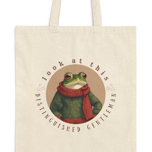 May include: A beige tote bag with a circular design featuring a green frog wearing a red scarf and a green sweater. The text around the frog reads "look at this distinguished gentleman".