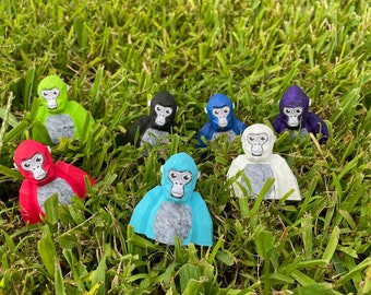 Puede incluir: Un grupo de ocho pequeñas figuritas de mono pintadas a mano en varios colores, incluyendo rojo, verde, azul, blanco, negro y morado. Las figuritas están dispuestas en círculo sobre un lecho de hierba verde.