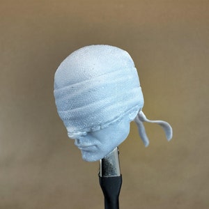 May include: A white 3D printed head with a blindfold. The blindfold is made of a textured fabric and is tied at the back of the head.