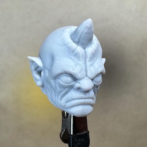 Pode incluir: Uma escultura cinza claro e detalhada de uma cabeça demoníaca com um único chifre, orelhas pontudas e uma expressão severa. A escultura é mantida no lugar por uma pinça de metal, sugerindo que é um trabalho em andamento ou um modelo.
