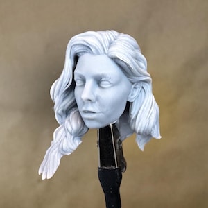 Pode incluir: Uma escultura azul clara da cabeça de uma mulher com cabelo longo e ondulado e uma trança. O rosto tem olhos fechados e uma expressão neutra. A escultura é montada em uma braçadeira preta contra um fundo bege.