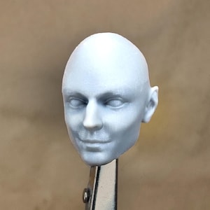 Op de afbeelding: Een wit, ongeverfd, plastic hoofd voor een pop of een action figure. Het hoofd heeft een neutrale uitdrukking met gesloten ogen en een licht geopende mond.