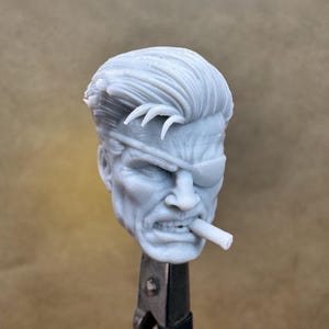 May include: A gray, detailed bust of a man with an eye patch, furrowed brow, and a cigar in his mouth. The sculpture features a stern expression and styled hair. The bust is mounted on a metal stand, suggesting it is a collectible or model.