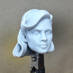 Puede incluir: Busto azul claro de una persona con cabello ondulado y expresión neutra. La escultura está sujeta por una pinza metálica, y el fondo es de color liso y neutro.