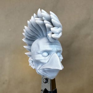 May include: A light gray sculpture of a stylized head with a prominent beak and a spiky, layered hairstyle. The sculpture is held in place by a metal clamp, suggesting it is a work in progress or a component of a larger piece.