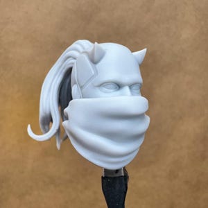 Puede incluir: Una cabeza esculpida de color gris claro con cuernos, una cubierta facial y cabello largo recogido en una cola de caballo. La cabeza está montada sobre una varilla negra, sobre un fondo beige liso.