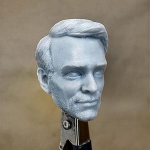 May include: A 3D printed head sculpt of a man with short, styled hair. The head is painted in a light gray color and has a realistic appearance.