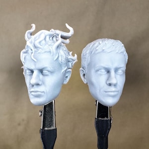 Puede incluir: Dos cabezas blancas de plástico sin pintar impresas en 3D, una con un peinado esculpido y la otra con un peinado más realista. Ambas cabezas están montadas en abrazaderas negras y plateadas.