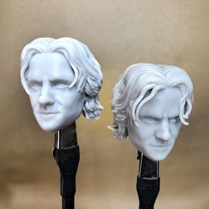 May include: Two gray resin busts of a man's head with wavy hair. The busts are mounted on black stands. The heads are facing forward with a neutral expression. The background is a solid tan color.
