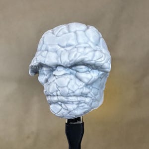 May include: A gray, textured, 3D printed model of a character's head. The character has a rocky, bumpy texture and a serious expression.
