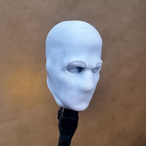 以下が含まれることがあります： 淡いグレーの3Dプリントされた人間の頭部の彫刻。滑らかで特徴のない顔立ち、窪んだ眼窩、質感のある表面が特徴です。頭部は黒い垂直の支柱に取り付けられており、未塗装の樹脂仕上げが施されています。