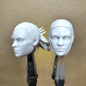 May include: Two unpainted, light gray resin head sculptures. One is in profile, showing a ponytail. The other is a frontal view, with a helmet-like structure. Both are mounted on metal clamps, suggesting they are works in progress.