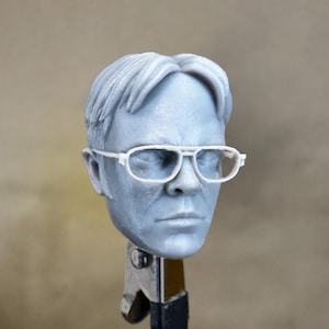 May include: A grey 3D printed head sculpt of a person wearing white framed glasses. The head is on a metal stand.