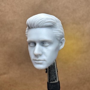 May include: A light gray, detailed sculpture of a human head with neatly styled hair. The sculpture features realistic facial features, including eyes, a nose, and a mouth. The head is held by a black clamp.