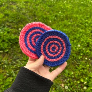 Op de afbeelding: Twee handgemaakte gehaakte onderzetters. Eén is roze met blauwe concentrische cirkels en de andere is blauw met roze concentrische cirkels. De onderzetters zijn rond en lijken van garen te zijn gemaakt. De achtergrond is wazig groen gras.