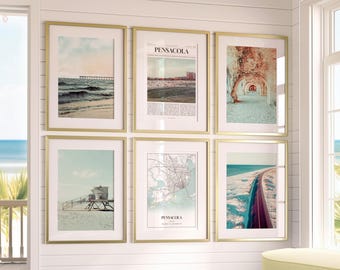May include: Six framed art prints with gold frames. The prints feature beach scenes, a map, and architectural images. The top center print has the word "PENSACOLA" in large text. The art is displayed on a white wall.