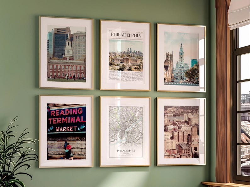K&ouml;nnte beinhalten: Sechs gerahmte Drucke mit goldfarbenen Rahmen h&auml;ngen an einer gr&uuml;nen Wand. Die Drucke zeigen Bilder von Philadelphia, darunter Stadtlandschaften, historische Geb&auml;ude und eine Karte. Ein Druck zeigt den Text "Reading Terminal Market."