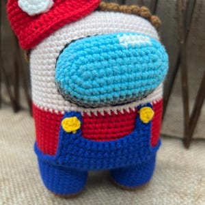 May include: A crocheted plush toy shaped like an Among Us character wearing a red hat and blue overalls. The toy has a white face with a blue visor and yellow buttons.