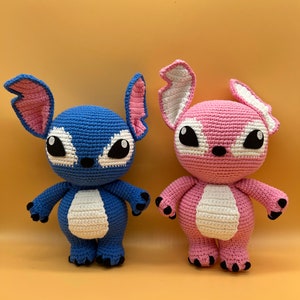 May include: Two crocheted plush toys, one blue and one pink, with white accents and black eyes. The toys are standing on a light yellow surface.