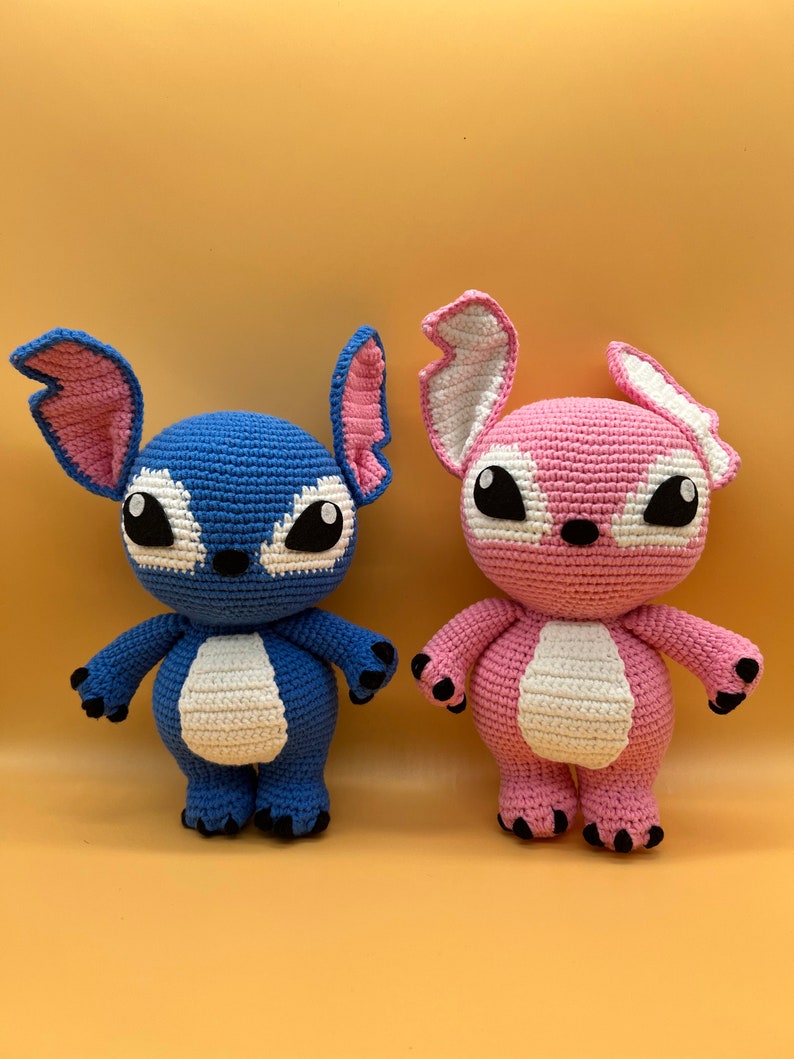 May include: Two crocheted plush toys, one blue and one pink, with white accents and black eyes. The toys are standing on a light yellow surface.