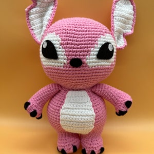 May include: Pink crocheted plush toy with large black eyes and white accents. The toy has a rounded body and long arms and legs.