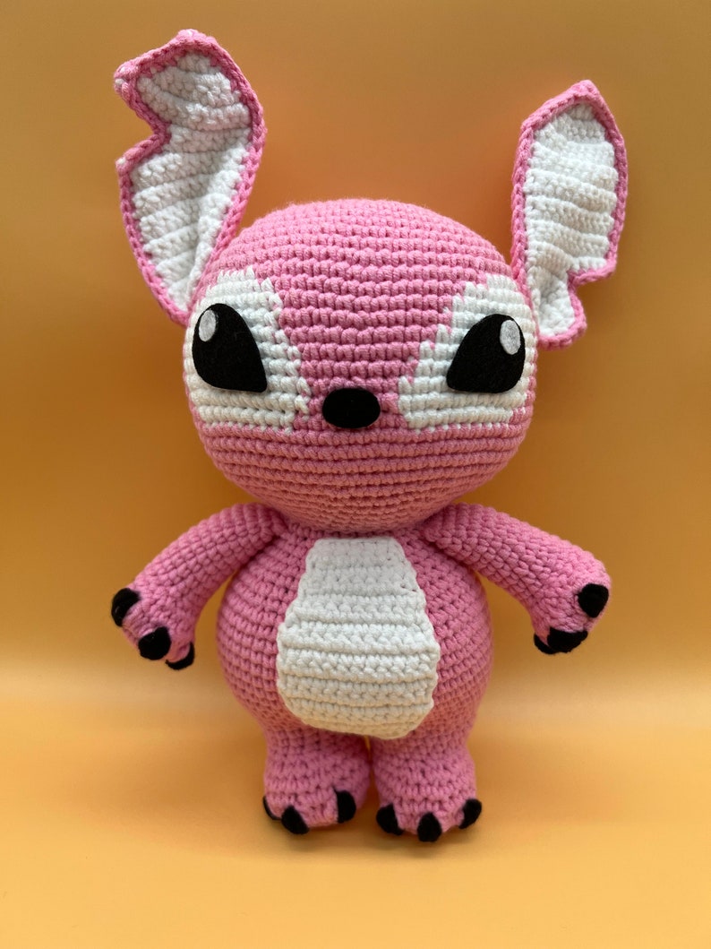 May include: Pink crocheted plush toy with large black eyes and white accents. The toy has a rounded body and long arms and legs.