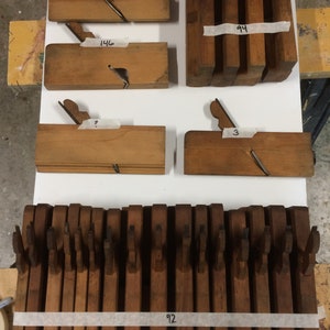 Op de afbeelding: Een verzameling houten handvlakken, sommige zijn in groepen gestapeld en andere liggen individueel. De vlakken zijn gelabeld met nummers, waaronder 92, 94, 146, 157 en 3.