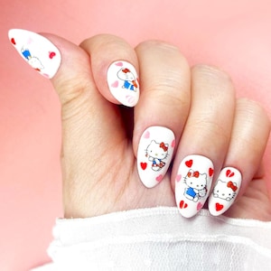 May include: White oval-shaped nail art featuring Hello Kitty designs. Each nail showcases a different Hello Kitty illustration, with red bows, hearts, and blue accents. The nails are painted with a glossy finish.
