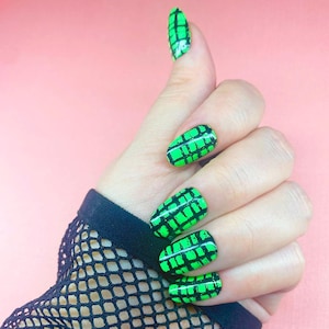 May include: A hand with green and black grid-patterned nail art. The nails are oval-shaped and painted with a bright green base and a black grid design. The background is a solid pink.