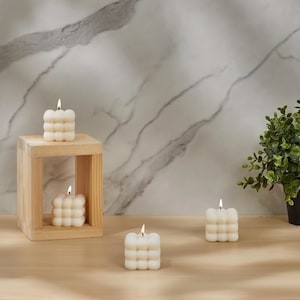 May include: Four white, round, square-shaped candles with a textured surface. The candles are lit and are on a wooden surface.