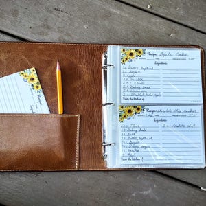 May include: A brown leather recipe binder with clear plastic sleeves. The binder has a pocket on the front cover and a pencil sticking out of the pocket. The sleeves contain recipe cards with handwritten recipes for apple cake and chocolate chip cookies.