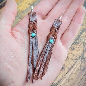 May include: Brown leather tassel earrings with turquoise stones. The earrings are braided and have a fringe detail.