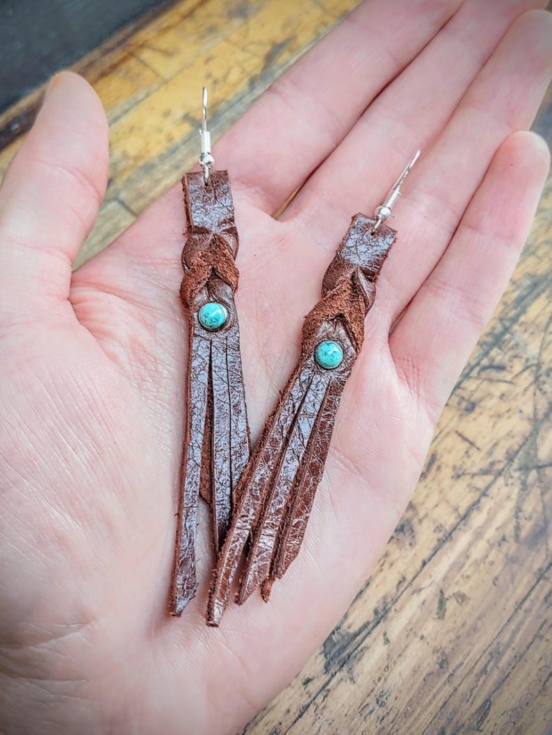 May include: Brown leather tassel earrings with turquoise stones. The earrings are braided and have a fringe detail.