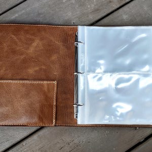 May include: A brown leather photo album with a clear plastic insert for holding photos. The album has a pocket on the front cover and a metal ring binder.