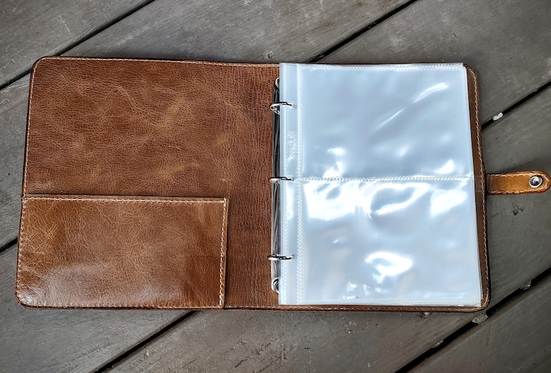 May include: A brown leather photo album with a clear plastic insert for holding photos. The album has a pocket on the front cover and a metal ring binder.
