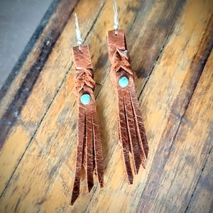 May include: A pair of brown leather tassel earrings with turquoise stones. The earrings are hanging from silver hooks.