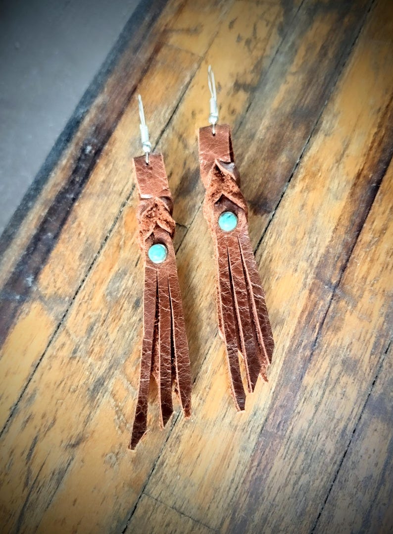 May include: A pair of brown leather tassel earrings with turquoise stones. The earrings are hanging from silver hooks.