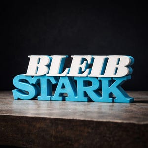 Può includere: Un cartello decorativo blu e bianco con le parole tedesche "BLEIB STARK" sovrapposte. Il cartello è composto da singole lettere e poggia su una superficie di legno scuro.