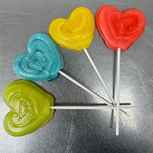 Puede incluir: Cuatro piruletas en forma de corazón en palos. Las piruletas son amarillas, azules, verdes y rojas.