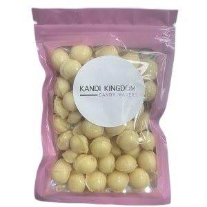 Può includere: Un sacchetto rosa richiudibile pieno di gocce di cioccolato bianco. Il sacchetto ha un'etichetta circolare bianca con il testo "KANDI KINGDOM CANDY MAKERS".